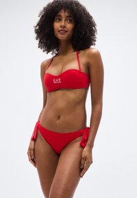 Σετ κόκκινου μπικίνι με τοπ bandeau που διαθέτει διακριτικό λογότυπο και σλιπ με δέσιμο στο πλάι. Λεπτό ύφασμα με εφαρμοστή γραμμή και μίνιμαλ στυλ.