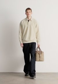 Krém színű, hosszú ujjú galléros póló logóval, sötét farmernadrággal párosítva; barna díszítéssel ellátott világosbarna táskát tartva. Az összhatást lapos cipő teszi teljessé.