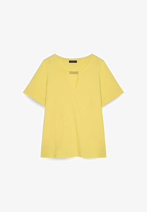 Żółta koszula z krótkim rękawem, okrągłym dekoltem i małym wycięciem w kształcie klucza zapinanym z przodu na srebrny dekoracyjny łańcuszek.