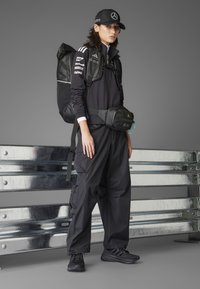 Schwarzes Sportoutfit mit einem Reißverschluss-Oberteil, lockeren Hosen, einem Rucksack und einer Bauchtasche, alles aus eleganten synthetischen Materialien. Schwarze Kappe mit Logo.