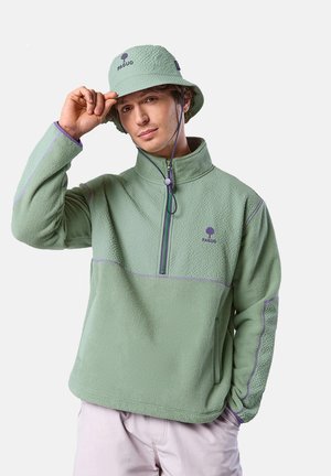 Giovane uomo che si sistema un cappello a secchiello verde menta, indossando un pile verde menta abbinato con cuciture viola e logo, in piedi contro uno sfondo bianco.