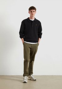 Polo shirt nero in maglia con colletto ripiegato, abbinato a pantaloni verdi oliva e sneakers beige. Presenta un piccolo emblema dorato sulla polo.