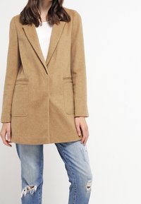 Béžové vlněné sako s jedním knoflíkem na zapínání, klopami s výstřihem a předními kapsami, ve spojení s potrhanými modrými džíny.