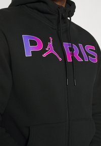 Sudadera con cremallera negra que presenta un gran texto "PARÍS" en un degradado de púrpura y rosa, con la silueta del logo de Air Jordan en el centro.