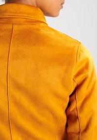 Gul fuskmockajacka med en slät yta, som har en klassisk krage och en figursydd form. Sömnadsdetaljer är synliga på baksidan.