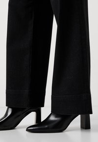 Pantalones negros de pierna ancha hechos de una tela texturizada, con un corte recto y sutiles destellos. Combinados con botas de tacón negro puntiagudas.