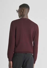 Maglione borgogna a trama con collo tondo e maniche lunghe, caratterizzato da un orlo e polsini a coste. Indossato con pantaloni neri su misura.