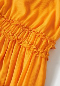 Tissu orange vif avec des sections froncées et des détails à volants, montrant la texture et les coutures sur un matériau léger et plissé.