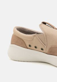 Beżowe sneakersy wsuwane z zamszowymi akcentami, trzema otworami wentylacyjnymi, elastycznym bocznym panelem i grubą białą amortyzowaną podeszwą na białym tle.