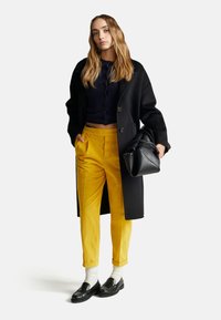 Zwarte jas over een marineblauw top, gele corduroybroek, witte sokken en zwarte instapschoenen. Houdt een gestructureerde zwarte handtas vast.