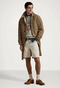 Muž ve světlehnědém kabátu, šedém svetru, modré košili, krémových šortkách, béžových ponožkách a hnědých botách, stojící s jednou rukou v kapse.