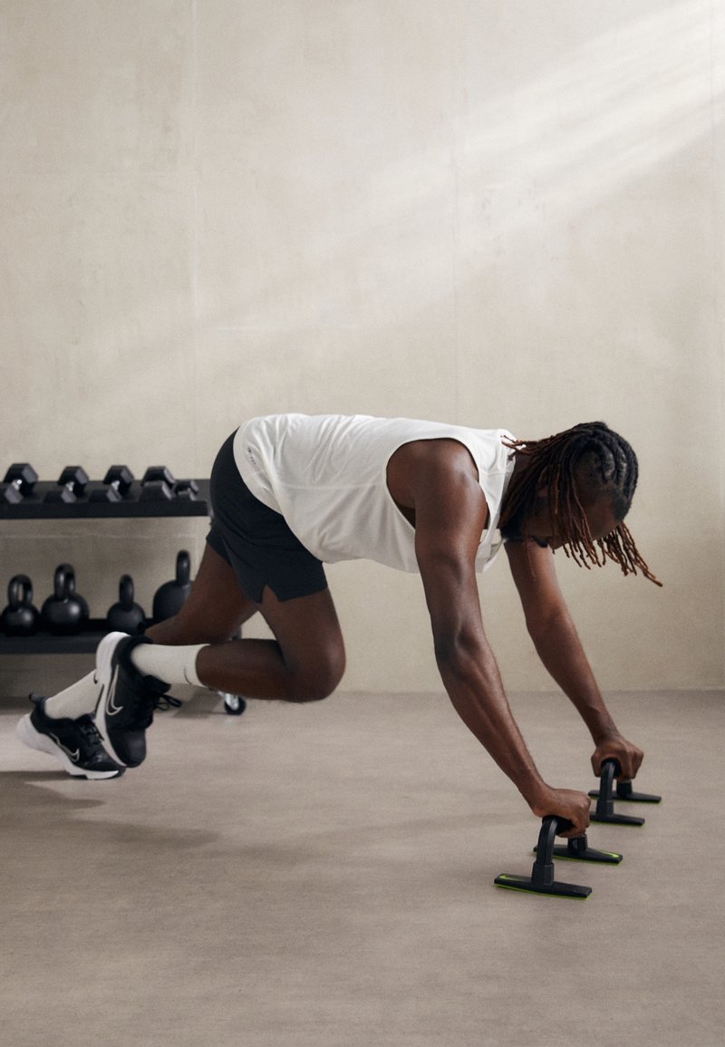 Homme en débardeur blanc et short noir faisant l'exercice du mountain climber avec des barres de pompe dans une salle de sport avec des poids en arrière-plan.