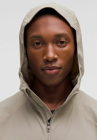 Giacca a vento beige chiaro con cappuccio e chiusura a zip, caratterizzata da una texture liscia e un design minimalista, indossata da un modello con un'espressione neutra.