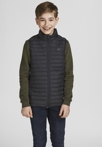 Marineblauwe puffervest met een hoge kraag, rits aan de voorkant en horizontale stiksels. Combinatie met een groene langemouwenshirt en blauwe spijkerbroek.