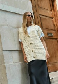 Cardigan en tricot crème avec un col en V, orné de grands boutons et d'un motif texturé, porté sur une jupe en satin noir.