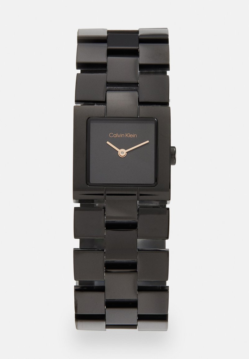 Must musta kell, millel on läikiv viimistlus, must dial, roosakuldne käed ja paks lingiga rihm. "Calvin Klein" on näidatud dialil.