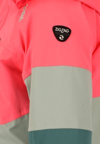 Farverig jakke med en lys pink øverste sektion, en lys grå midtersektion og en mørkegrøn nederste sektion. Viser et sort logo-patch.