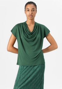 Top verde drappeggiato con maniche corte, realizzato in un tessuto morbido, con scollo a cowl. Abbinato a una gonna plissettata verde metallica.
