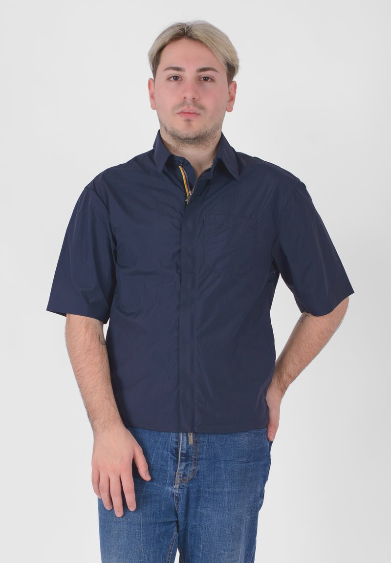 Camicia a maniche corte, di colore blu navy, realizzata in materiale leggero. Presenta una tasca frontale e un colletto con zip, arricchito da dettagli colorati.