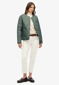 Gequilted groene jas met ronde halslijn en drukknoopsluiting, gedragen over een crèmekleurige turtleneck, gecombineerd met witte broek en donkere loafers.