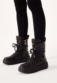 Schwarze, gepolsterte Knöchelstiefel mit einer dicken, geriffelten Sohle, Schnürverschluss vorne, seitlichem Reißverschluss und metallischen Hardware-Akzenten. Strukturmaterial, robust und langlebig.