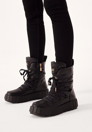 Bottes de cheville noires duveteuses avec une épaisse semelle crantée, un laçage à l'avant, une fermeture éclair sur le côté et des accents en métal. Matériau texturé et durable.