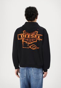 Čierna mikina s kapucňou s veľkým oranžovým potlačou "DIESEL" na chrbte, ktorá má text�úrovaný dizajn. Má uvoľnený strih a klasickú klokankovú kapsu.