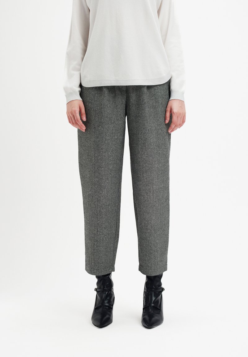 Osoba ubrana w szare spodnie w jodełkę, białą bluzkę z długim rękawem oraz czarne botki za kostkę stoi na białym tle.