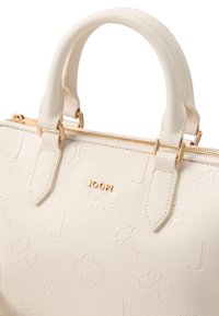 Sac à main texturé blanc avec quincaillerie dorée, motifs floraux et lettres en relief, double poignées et fermeture zippée sur le dessus. Logo affiché de manière proéminente.