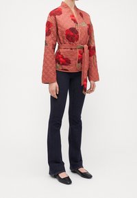 Vatteret, lyserød jakke med stort rødt blomstermønster, bælte i taljen, lange ærmer, båret over en mørk bluse og blå jeans, parret med sorte flade sko.
