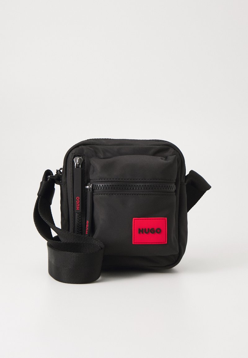 Borsa nera in nylon dalla forma rettangolare, con una patch rossa con logo sulla parte frontale, due cerniere e una tracolla regolabile.