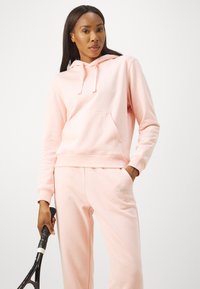 Felpa rosa chiaro con tasca a marsupio e cordoncino, abbinata a pantaloni della tuta coordinati. Entrambi presentano un morbido materiale felpato e un branding discreto.