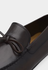 Κοντινό πλάνο ενός καφέ σκούρου δερμάτινου μοκασίνι με διακοσμητικό δερμάτινο φιόγκο στο επάνω μέρος και εμφανή ραφή κατά μήκος των άκρων.