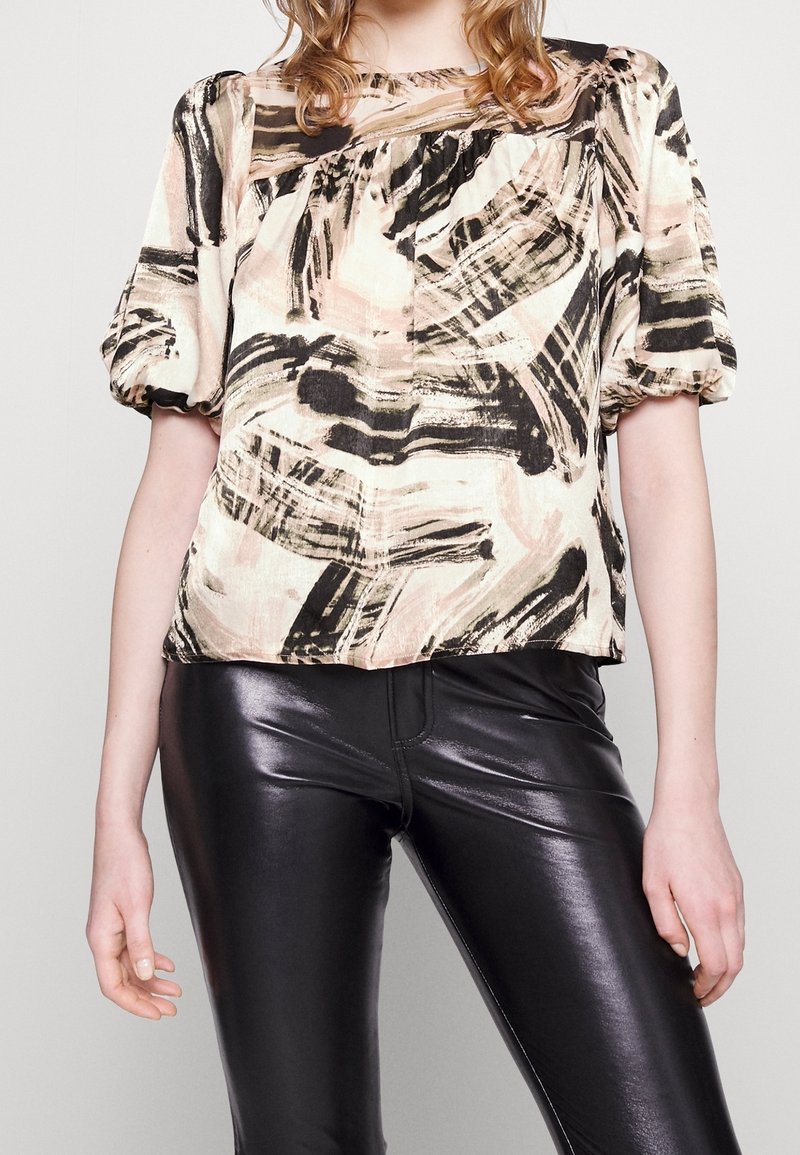 Blusa a fantasia floreale con maniche a sbuffo, realizzata in un tessuto leggero nei toni beige, nero e verde, indossata con leggings neri lucidi.