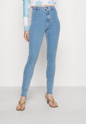 Nainen, jolla on vaaleansiniset kapealahkeiset farkut ja beiget korolliset sandaalit, seisoo yksiväristä valkoista taustaa vasten.