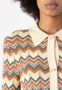 Strikket cardigan med en krave og knap i cremefarve, med et zigzag-mønster i pink, grå og sennepsgul. Tekstureret stof.