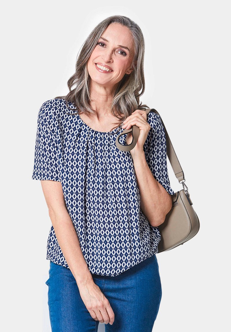 Marineblauwe blouse met een patroon van ruiten, korte mouwen en een ronde hals; gecombineerd met blauwe jeans en een lichtbeige handtas.