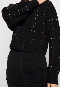 Cardigan noir avec texture côtelée, featuring perles synthétiques colorées en rose et violet. Design court avec fermetures à boutons.