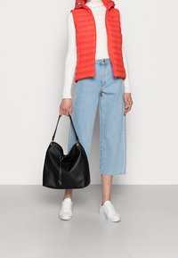 Svart läderhobo-väska med mjuk textur, böjd form och guldaccenter. Modellen bär en röd puffväst, vit tröja och ljusblå cropped jeans.