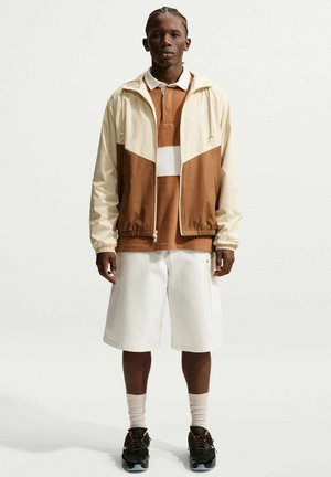 Joven de pie con chaqueta marrón y blanca, camisa marrón, pantalones cortos blancos, calcetines beige y zapatillas negras sobre un fondo liso.
