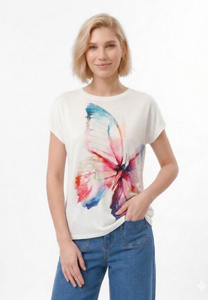 Γυναίκα με κοντά ξανθά μαλλιά που φοράει λευκό μπλουζάκι με μια μεγάλη πολύχρωμη πεταλούδα σε σχέδιο υδατογραφίας και μπλε τζιν, χαμογελώντας και έχοντας το ένα χέρι στην τσέπη.