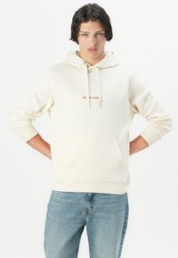 Crème hoodie met een kangaroo pocket, ribgebreide manchetten en zoom. Kenmerkt zich door een klein bruin geborduurd logo op de borst. Draagt met een blauwe jeans.