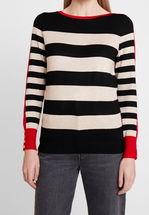Femme portant un pull en maille à manches longues avec de larges rayures horizontales noires et beiges et des poignets rouges avec des boutons, assorti à un jean gris.
