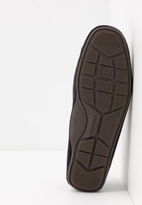 Semelle de chaussure noire avec une semelle extérieure en caoutchouc marron à motif et "HILFIGER" embossé au centre, incliné contre un mur blanc d'angle.