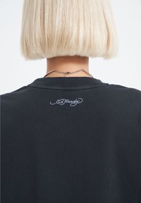 Černé tričko s kulatým výstřihem, na zadní straně s bílou vyšívanou logem "Ed Hardy". Měkká tkanina s hladkou texturou.