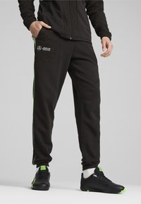 Zwarte trainingsbroek met een gestructureerd paneel en groene accenten, voorzien van zijzakken en nauwsluitende manchetten. Gecombineerd met zwarte sportschoenen.