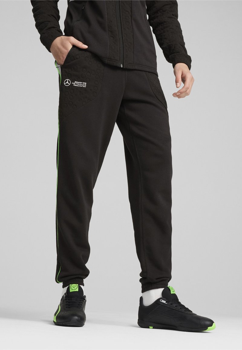 Zwarte trainingsbroek met een gestructureerd paneel en groene accenten, voorzien van zijzakken en nauwsluitende manchetten. Gecombineerd met zwarte sportschoenen.