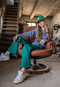 Groene wijde broek, spijkerjack en witte sneakers. Zittend in een bruine leren draaistoel in een rustieke inrichting met houten balken.