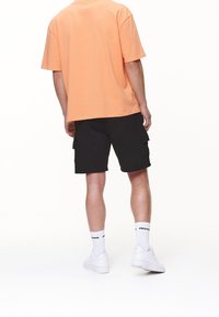 Hombre vestido con una camiseta de color durazno, pantalones cortos cargo negros, zapatillas Nike Air blancas y calcetines blancos con texto negro "Pegador", de espaldas a la cámara.