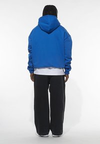 Modrá mikina s kapucí s volným střihem, žebrovanými manžetami a lemem. Nošená přes bílou vrstvu košile, doplněná volnými černými kalhotami.